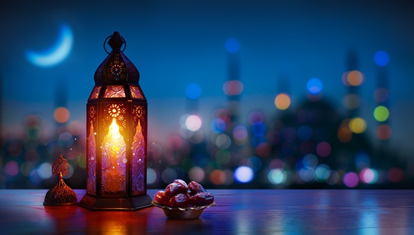 A glowing lantern beside a bowl of dates with a blurred city and crescent moon in the background.