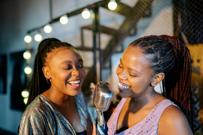 Two people sing into a vintage microphone at a karaoke venue with string lights in the background.